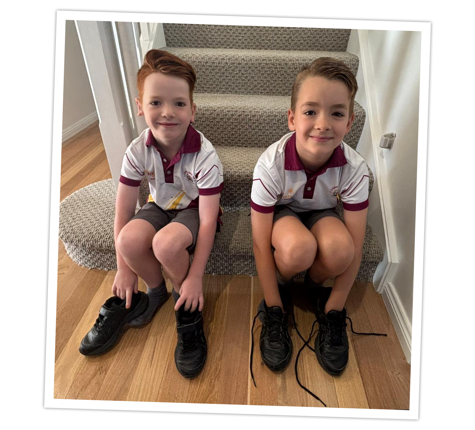 Two children sitting on a carpeted staircase indoors, wearing matching school uniforms with white shirts featuring maroon collars and grey shorts. Each child is holding or wearing black school shoes, with one pair untied and laces visible on the floor. The setting includes wooden flooring and a neutral-colored wall.