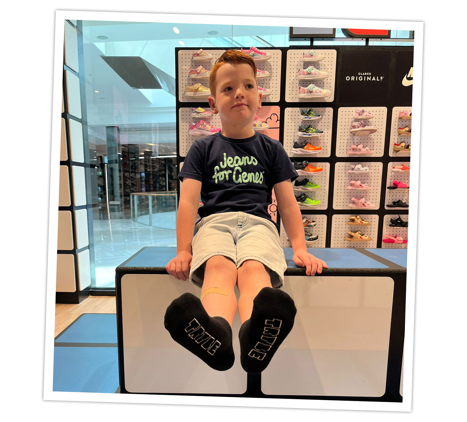 A child sitting on a padded bench inside a shoe store, wearing a dark blue “Jeans for Genes” t-shirt, light-colored shorts, and black socks with white text that reads “TIME.” Behind the child is a display wall filled with colorful athletic shoes arranged in rows. The setting appears to be a modern retail environment with bright lighting and glass panels.